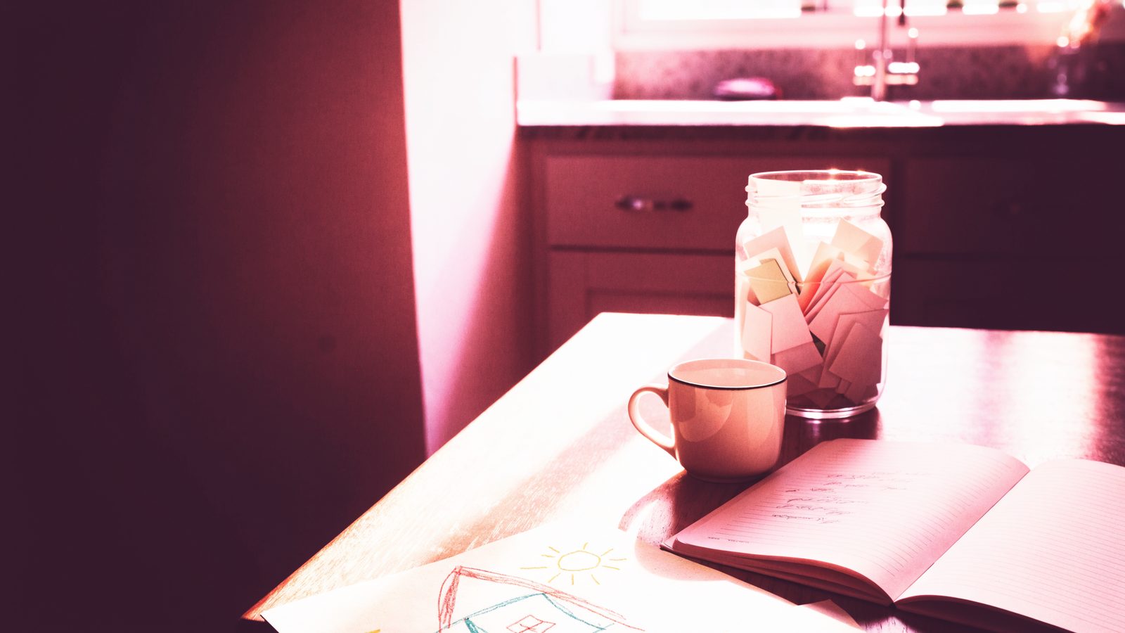 A sunlit kitchen table with an open notebook, a coffee mug and a glass jar filled with small folded paper notes, in soft pink morning light.