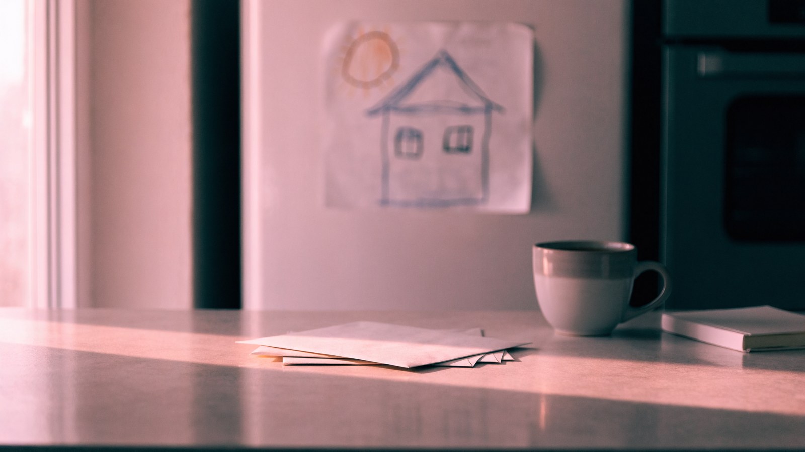 A kitchen counter in soft pink morning light with a stack of envelopes, a coffee mug and a small notebook, with a child's drawing on the fridge behind.