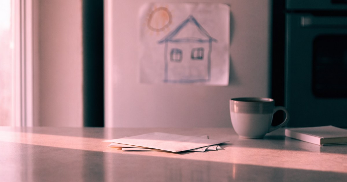 A kitchen counter in soft pink morning light with a stack of envelopes, a coffee mug and a small notebook, with a child's drawing on the fridge behind.